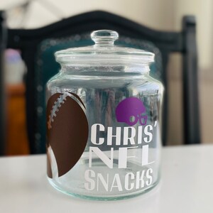 May include: A clear glass jar with a lid, decorated with a brown football, a purple football helmet, and the text "Chris' NFL Snacks".