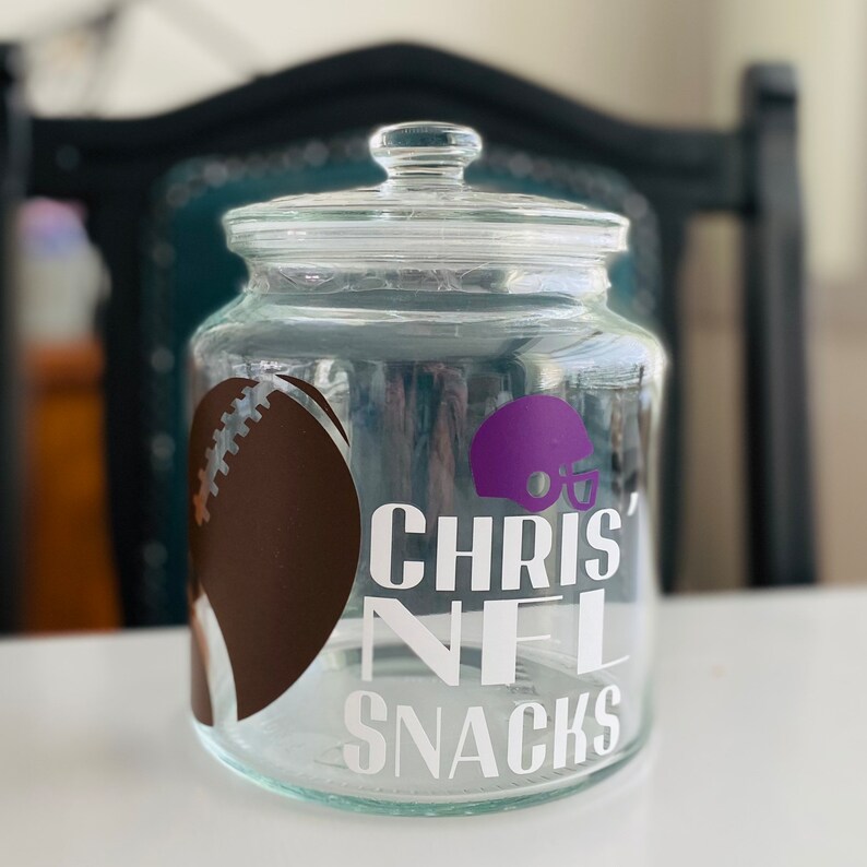 May include: A clear glass jar with a lid, decorated with a brown football, a purple football helmet, and the text "Chris' NFL Snacks".