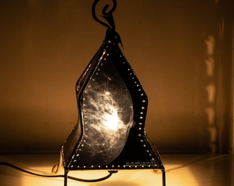 Puede incluir: Un farol de metal negro con parte superior puntiaguda y patas curvas, con paneles translúcidos que muestran una imagen de mamografía médica en escala de grises. El farol emite una luz cálida y brillante, proyectando un reflejo nítido.