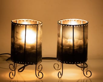 Puede incluir: Un par de lámparas de mesa cilíndricas con paneles de vidrio ahumado oscuro enmarcados por finas tiras de metal. Cada lámpara descansa sobre tres patas de metal decorativas y curvadas, emitiendo una luz cálida y brillante desde su interior.
