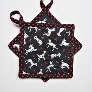 May include: Two square black pot holders with a Dalmatian dog pattern and red heart trim. The pot holders have a quilted design and a loop for hanging. The dogs are white with black spots.