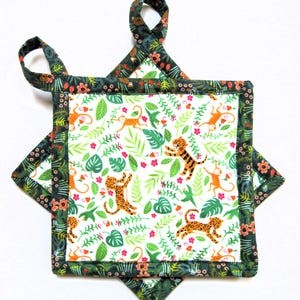 May include: A quilted pot holder with a jungle theme. The center features a white background with orange monkeys, tigers, and green leaves. The edges are dark green with a floral pattern. Two loops are at the top for hanging.
