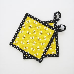 May include: Two square quilted pot holders with a yellow background and a pattern of bees. The pot holders have a black polka dot border and a loop for hanging. The pot holders are approximately 8 inches square.