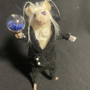 May include: A whimsical, handcrafted mouse figurine dressed in a black suit with white buttons and a ruffled collar. The mouse holds a glass orb filled with blue crystals. The mouse has white hair and purple eye makeup.