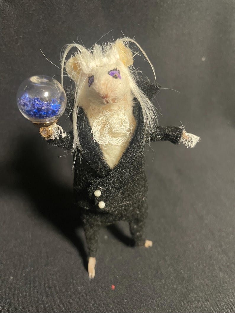 May include: A whimsical, handcrafted mouse figurine dressed in a black suit with white buttons and a ruffled collar. The mouse holds a glass orb filled with blue crystals. The mouse has white hair and purple eye makeup.