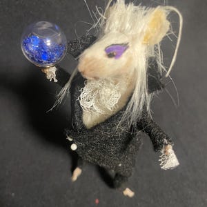 May include: A whimsical miniature mouse figure dressed in a black coat with a white lace collar and cuffs. The mouse has white fur, purple eye makeup, and holds a small glass orb filled with blue glitter. The figure is on a dark surface.