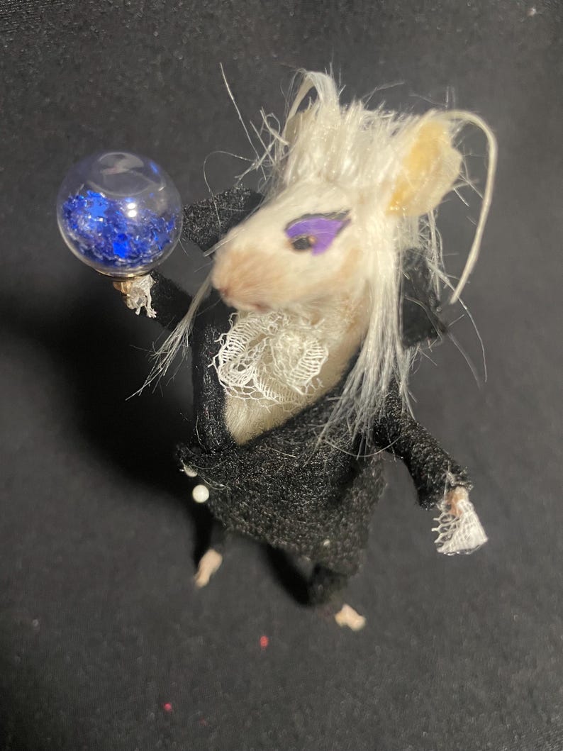 May include: A whimsical miniature mouse figure dressed in a black coat with a white lace collar and cuffs. The mouse has white fur, purple eye makeup, and holds a small glass orb filled with blue glitter. The figure is on a dark surface.