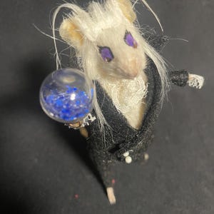 May include: A miniature mouse figurine with white fur and purple eyes, dressed in a black suit with a white lace collar. The mouse holds a clear orb filled with blue glitter, creating a whimsical and detailed aesthetic.