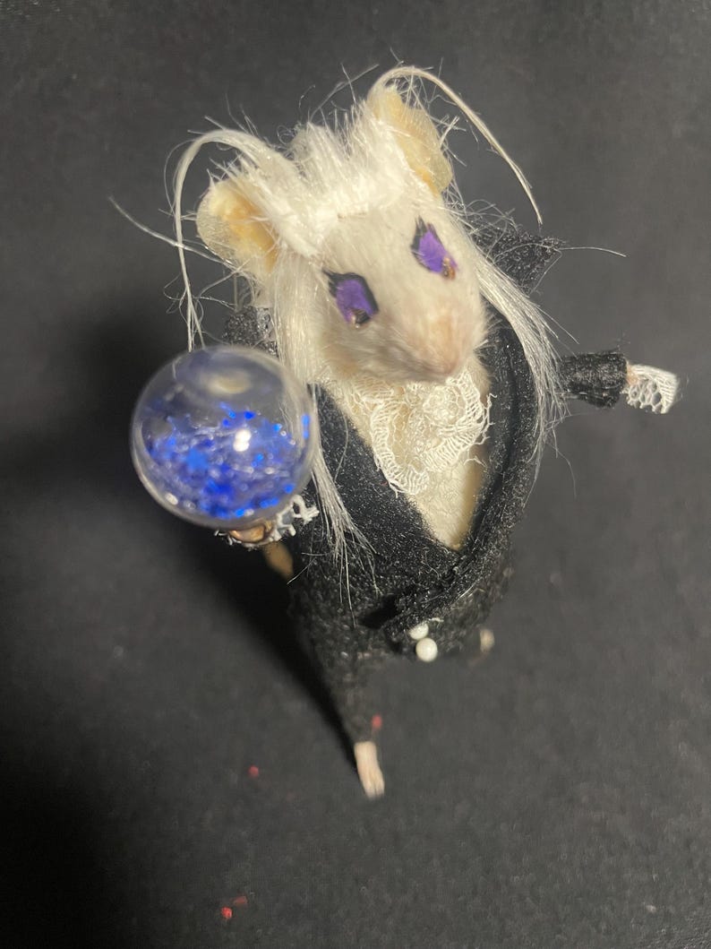 May include: A miniature mouse figurine with white fur and purple eyes, dressed in a black suit with a white lace collar. The mouse holds a clear orb filled with blue glitter, creating a whimsical and detailed aesthetic.