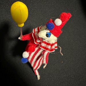 Może przedstawiać: Kapryśna, zabytkowa figurka myszy-klauna. Mysz ubrana jest w czerwono-biały pasiasty str&oacute;j z pasującym kapeluszem ozdobionym pomponami. Trzyma ż&oacute;łty balon, tworząc zabawną i nostalgiczną estetykę.