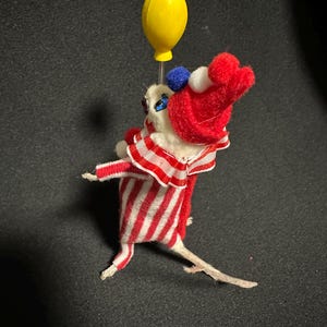 Może przedstawiać: Kapryśna, ręcznie robiona figurka myszy przebrana za klauna, trzymająca ż&oacute;łty balon. Mysz nosi czerwono-biały pasiasty str&oacute;j z pasującym kapeluszem i marszczonym kołnierzem. Figurka ma małe niebieskie i białe akcenty.