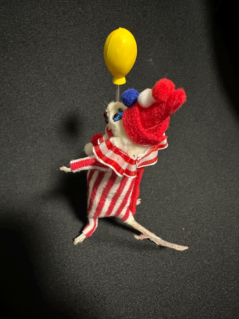 Może przedstawiać: Kapryśna, ręcznie robiona figurka myszy przebrana za klauna, trzymająca ż&oacute;łty balon. Mysz nosi czerwono-biały pasiasty str&oacute;j z pasującym kapeluszem i marszczonym kołnierzem. Figurka ma małe niebieskie i białe akcenty.