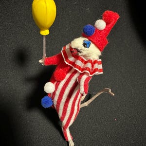 Może przedstawiać: Figurka klauna w stylu vintage. Klaun ubrany jest w czerwono-biały pasiasty str&oacute;j i pasujący kapelusz z pomponami. Trzyma ż&oacute;łty balon. Twarz klauna jest biała z niebieskimi oczami i czarnymi wąsami.