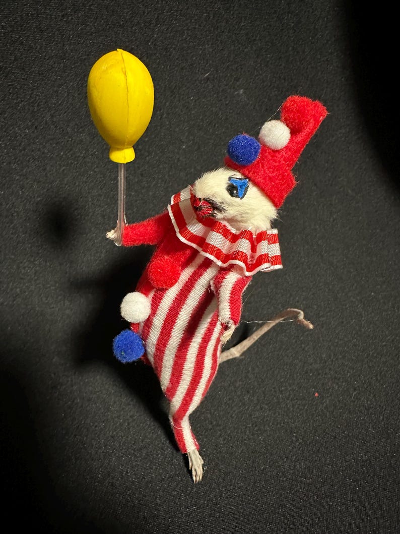 Może przedstawiać: Figurka klauna w stylu vintage. Klaun ubrany jest w czerwono-biały pasiasty str&oacute;j i pasujący kapelusz z pomponami. Trzyma ż&oacute;łty balon. Twarz klauna jest biała z niebieskimi oczami i czarnymi wąsami.