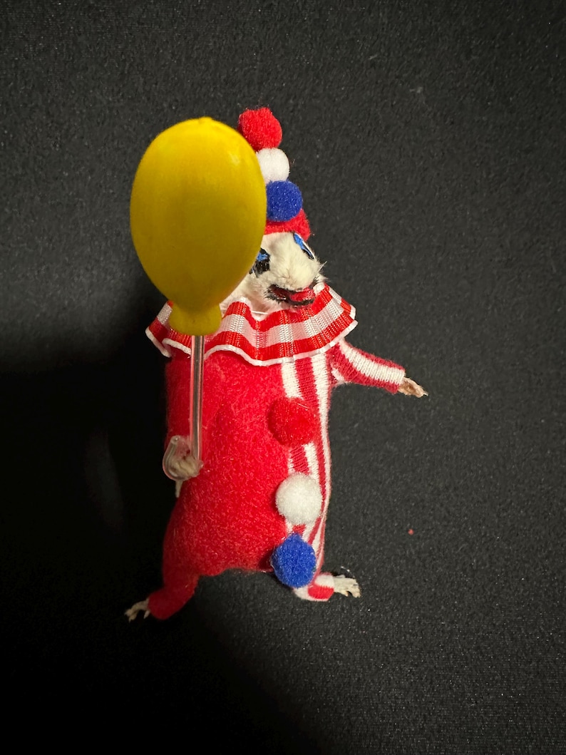 Może przedstawiać: Kapryśna, ręcznie robiona figurka klauna. Czerwono-biały pasiasty str&oacute;j ma kolorowe pompony i marszczony kołnierz. Klaun trzyma ż&oacute;łty balon. Kapelusz klauna jest czerwony, biały i niebieski.