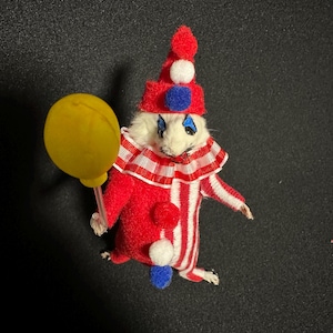 Może przedstawiać: Miniaturowa figurka klauna w czerwono-białym pasiastym stroju, czerwonym kapeluszu z białymi i niebieskimi detalami oraz ż&oacute;łtym balonem. Klaun ma białą twarz z niebieskim makijażem oczu. Fantazyjny przedmiot dekoracyjny.