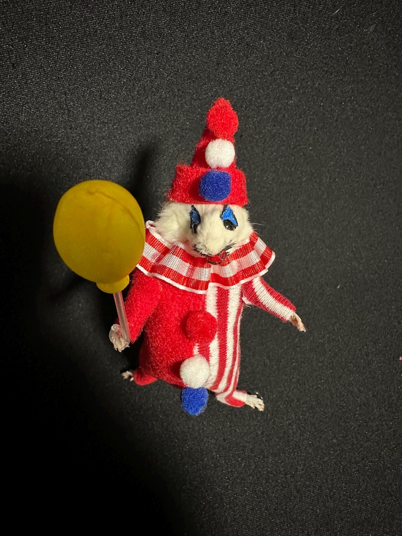 Może przedstawiać: Miniaturowa figurka klauna w czerwono-białym pasiastym stroju, czerwonym kapeluszu z białymi i niebieskimi detalami oraz ż&oacute;łtym balonem. Klaun ma białą twarz z niebieskim makijażem oczu. Fantazyjny przedmiot dekoracyjny.