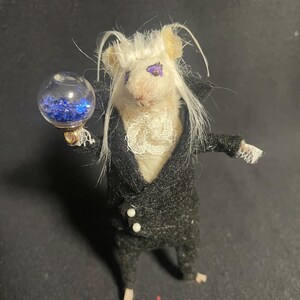 May include: A miniature mouse figure dressed in a black suit with white trim and a ruffled collar. The mouse has white hair and purple eyes, and holds a clear orb filled with blue glitter. The figure is posed against a dark background.