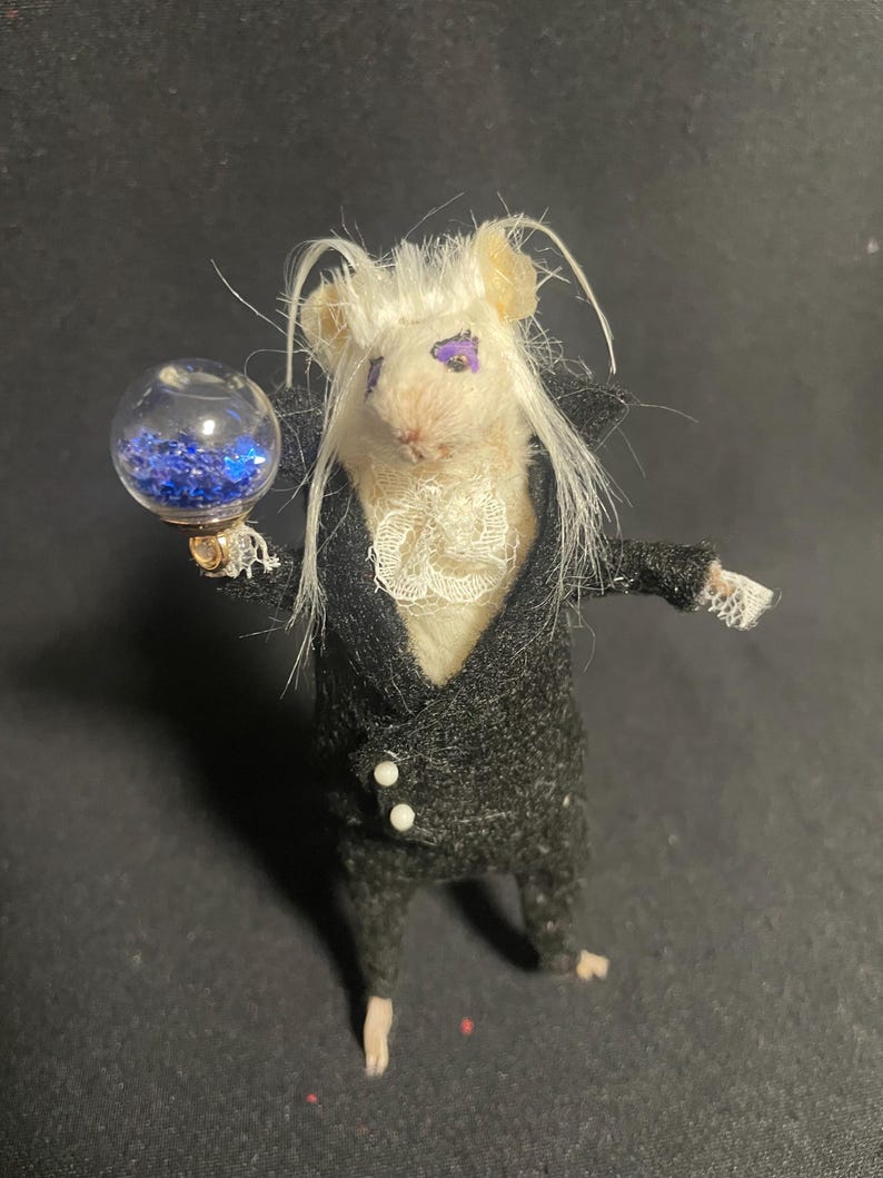 May include: A miniature mouse figure dressed in a black suit with white trim and a ruffled collar. The mouse has white hair and purple eyes, and holds a clear orb filled with blue glitter. The figure is posed against a dark background.