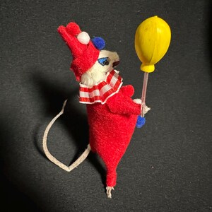 Może przedstawiać: Figurka myszy w stylu vintage przebrana za klauna, w czerwonym stroju, biało-czerwonym kołnierzu w paski i czerwonym kapeluszu z białymi i niebieskimi detalami. Trzyma ż&oacute;łty balon.