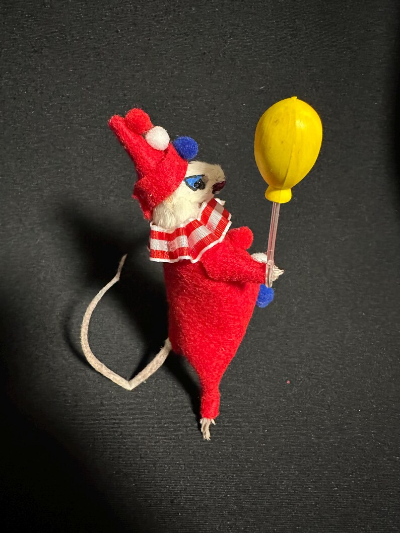 Może przedstawiać: Figurka myszy w stylu vintage przebrana za klauna, w czerwonym stroju, biało-czerwonym kołnierzu w paski i czerwonym kapeluszu z białymi i niebieskimi detalami. Trzyma ż&oacute;łty balon.