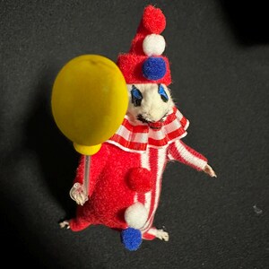Może przedstawiać: Figurka klauna w stylu vintage. Klaun ma na sobie czerwono-biały pasiasty str&oacute;j i trzyma ż&oacute;łty balon. Ma czerwony kapelusz z białymi i niebieskimi pomponami oraz białą twarz z namalowanymi cechami. Przedmiot kolekcjonerski.