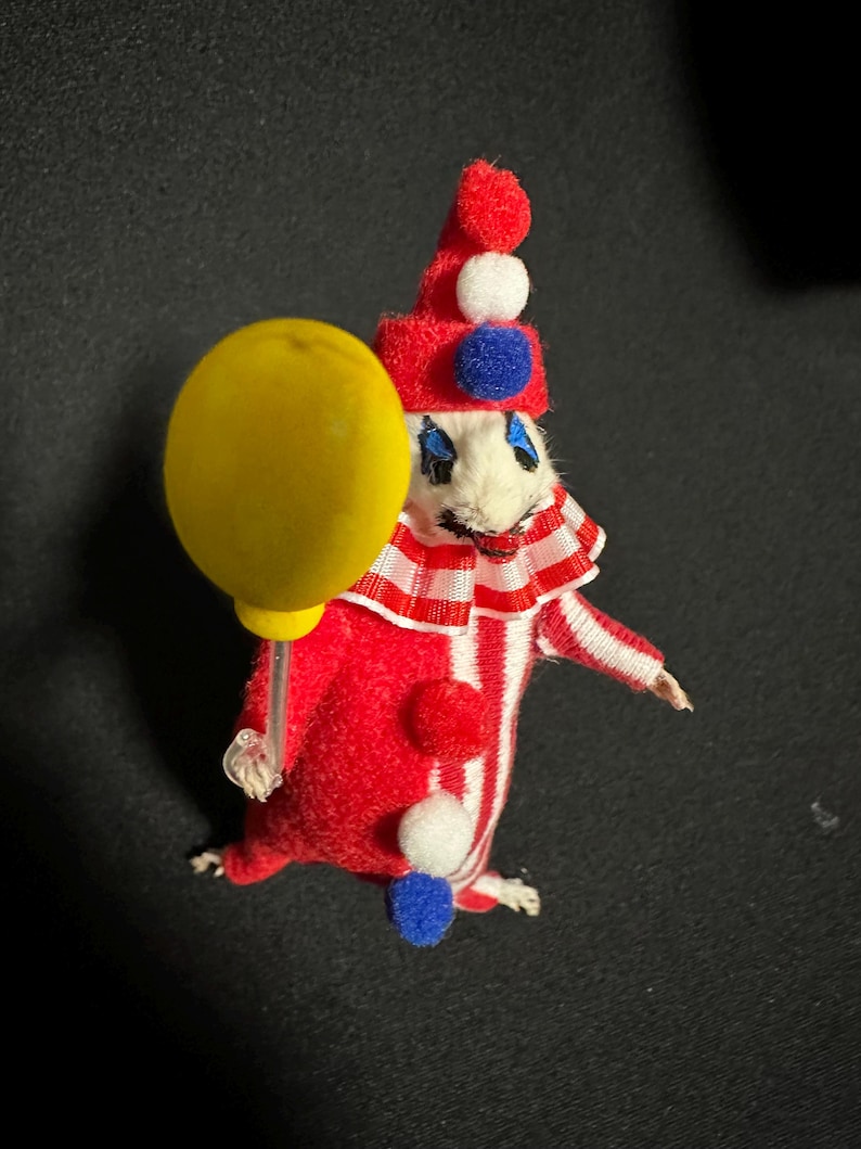 Może przedstawiać: Figurka klauna w stylu vintage. Klaun ma na sobie czerwono-biały pasiasty str&oacute;j i trzyma ż&oacute;łty balon. Ma czerwony kapelusz z białymi i niebieskimi pomponami oraz białą twarz z namalowanymi cechami. Przedmiot kolekcjonerski.