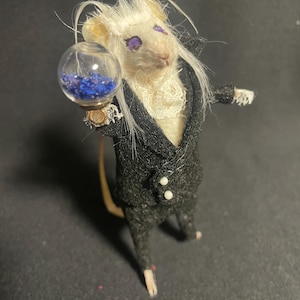 May include: A miniature mouse figure wearing a black suit with a white ruffled shirt and holding a glass orb filled with blue beads. The mouse has white fur, purple eyes, and a long tail.