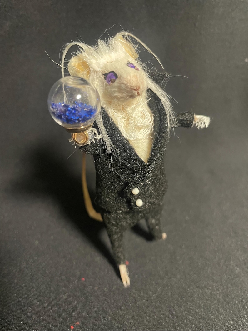 May include: A miniature mouse figure wearing a black suit with a white ruffled shirt and holding a glass orb filled with blue beads. The mouse has white fur, purple eyes, and a long tail.