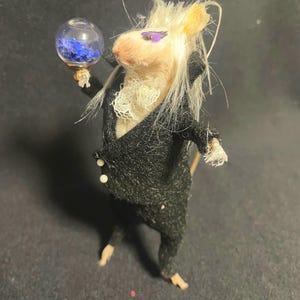May include: A whimsical, handmade mouse figurine dressed in a black suit with white accents. The mouse holds a small glass orb filled with blue material. The mouse has a cream-colored face, purple eye makeup, and long, white hair.