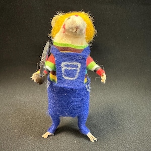 May include: A handmade felted mouse figurine wearing a blue jumpsuit with a white pocket, a striped shirt, and a yellow wig. The mouse holds a small knife. The figurine is set against a dark background.