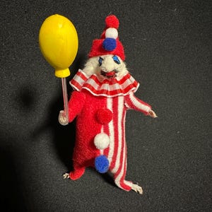 Może przedstawiać: Kapryśna figurka klauna z czerwono-białym pasiastym korpusem, trzymająca ż&oacute;łty balon. Klaun ma czerwony kapelusz z pomponami i marszczony kołnierz. Unikalny przedmiot dekoracyjny.