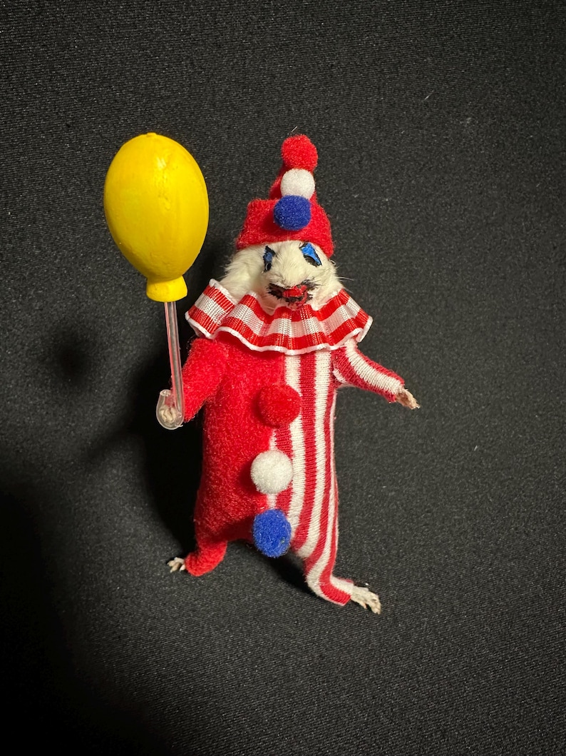Może przedstawiać: Kapryśna figurka klauna z czerwono-białym pasiastym korpusem, trzymająca ż&oacute;łty balon. Klaun ma czerwony kapelusz z pomponami i marszczony kołnierz. Unikalny przedmiot dekoracyjny.