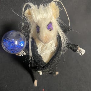 May include: A miniature mouse figure with white fur and long white hair, wearing a black suit with a white lace collar. The mouse has purple eyes and is holding a clear orb filled with blue glitter. The figure is a whimsical, fantasy-themed creation.