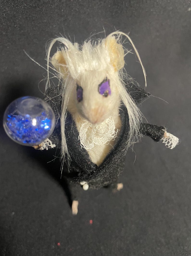 May include: A miniature mouse figure with white fur and long white hair, wearing a black suit with a white lace collar. The mouse has purple eyes and is holding a clear orb filled with blue glitter. The figure is a whimsical, fantasy-themed creation.