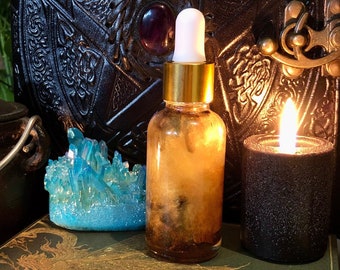 May include: A glass bottle with a gold lid and a dropper filled with a brown liquid. The bottle is sitting on a stack of books with a blue crystal and a black candle in the background.
