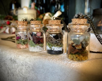 May include: Four small glass bottles with cork tops and a variety of natural materials inside, including crystals, herbs, and stones. The bottles are on a white surface and are attached to a bronze chain.