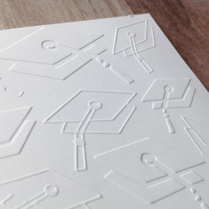 May include: White card with a repeating embossed pattern of graduation caps. The caps are outlined in white and have a tassel at the top.