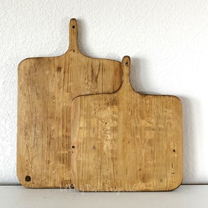 May include: Two wooden cutting boards with handles. The boards are rectangular with rounded corners and a natural wood grain finish. The boards are different sizes, with the larger one in front and the smaller one behind it. The boards are light brown in color.
