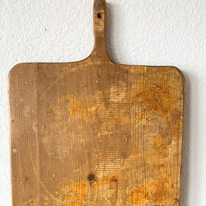 May include: A large, rectangular wooden cutting board with a handle. The board has a natural wood finish and shows signs of use, with orange stains and marks.