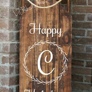 May include: A rustic wooden sled-shaped sign with the words "Happy Holidays" in white script. The sign features a white wreath around a large white "C". A rope is attached to the top for hanging.