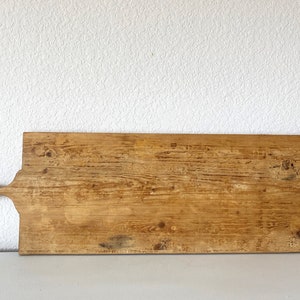 May include: A rectangular wooden cutting board with a handle. The board is made of light-colored wood with visible grain and knots. The handle has a hole for hanging. The board is approximately 24 inches long.