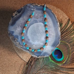 Op de afbeelding: Een turquoise en bruine kralenketting met een zilveren sluiting. De ketting wordt getoond op een grijze en witte agaat plak.