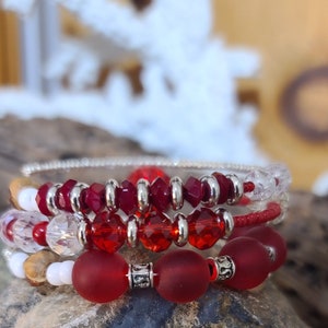 May include: Three beaded bracelets in shades of red, white, and clear. The bracelets feature various bead styles, including faceted, round, and cylindrical, with silver-toned metal accents. The bracelets are stacked together, creating a layered look.