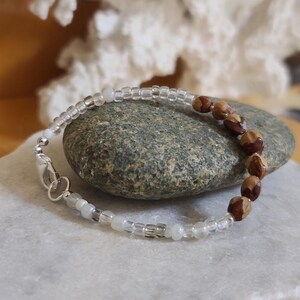 May include: A beaded bracelet with a mix of clear and brown beads. The bracelet is displayed on a white marble surface, with a gray stone and white coral in the background. The bracelet has a silver clasp.