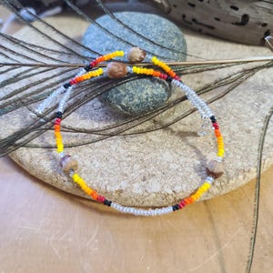 May include: A beaded bracelet with a colourful design. The bracelet features small, round beads in shades of yellow, orange, red, black, and silver. Three brown, organic-shaped beads are spaced along the bracelet. The bracelet is displayed on a textured, light brown surface.