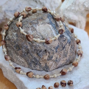 May include: A double strand necklace made of natural brown and white beads. The necklace is displayed on a rock and a white surface with loose beads scattered around.