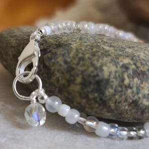 May include: A delicate bracelet featuring iridescent white beads and a silver clasp. A small, clear charm with a faceted design hangs from the clasp. The bracelet is displayed on a textured stone surface.