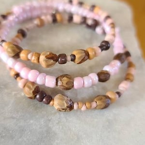 Op de afbeelding: Drie gelaagde armbanden met roze, bruine en natuurlijke houten kralen. De armbanden liggen op een wit oppervlak.