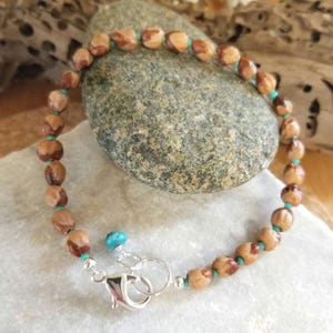 May include: A bracelet made of brown, oblong beads and small turquoise beads. The bracelet has a silver clasp and a small turquoise accent bead. The bracelet is displayed on a stone surface.